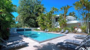 A swimming pool with lounge chairs, surrounded by tropical vegetation and a white building.
