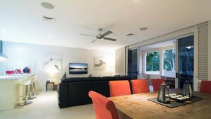 A modern living room with a wooden dining table, orange chairs, a black sofa, and a ceiling fan.