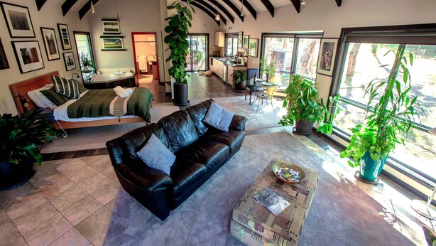 Amplia sala de estar con un sofá de cuero negro, una mesa de centro y una planta grande en maceta. Se observa una cama con colcha verde y varias almohadas. El ambiente cuenta con techos altos con vigas a la vista y ventanas grandes que ofrecen luz natural.