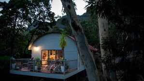 Una casa moderna de una sola planta con techo curvo, rodeada de árboles. Cuenta con un balcón con mobiliario exterior y un ventanal grande.