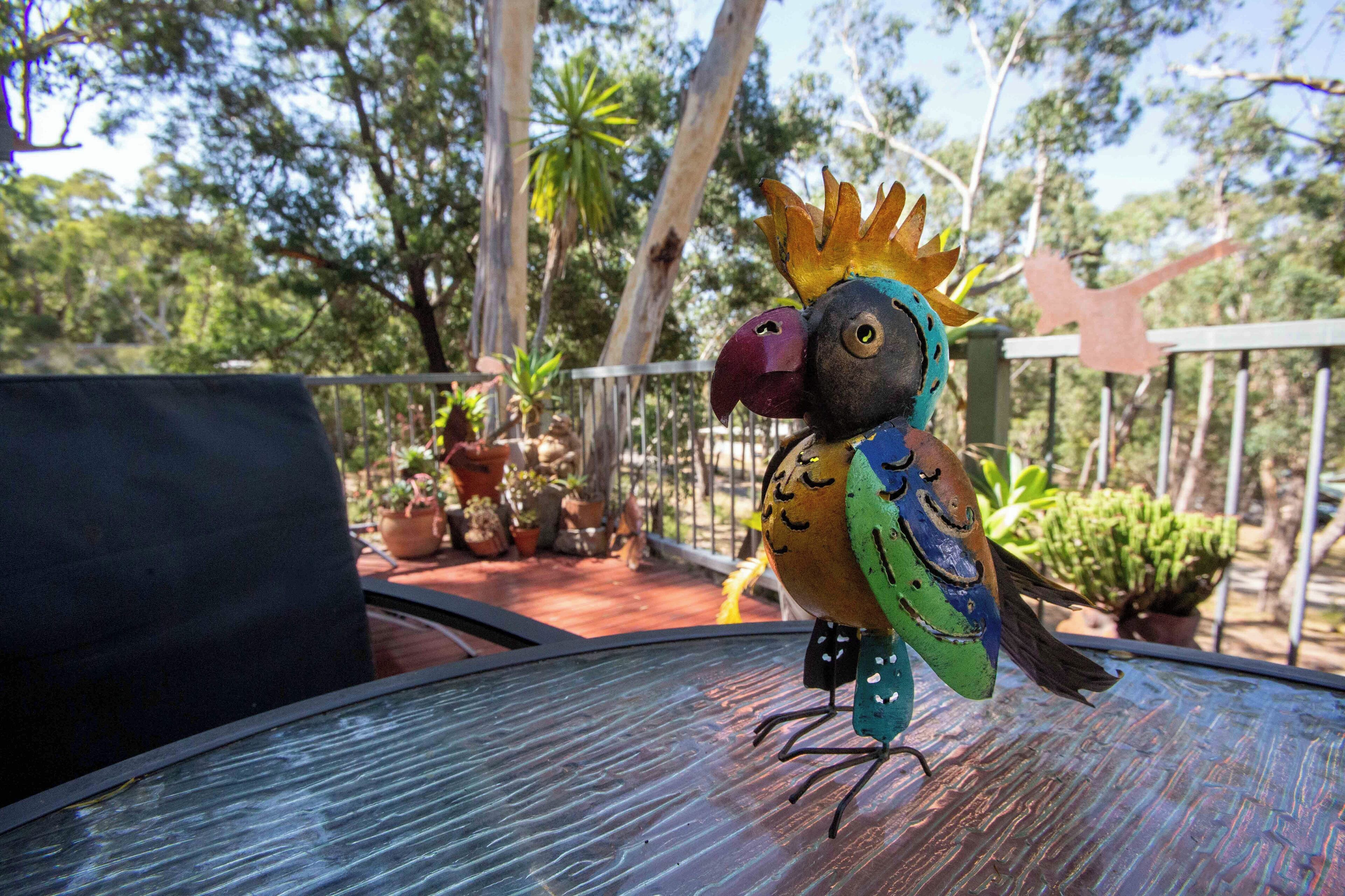 Une sculpture d’oiseau aux couleurs vives, posée sur une table en verre à l’extérieur, avec des plantes en pot et des arbres en arrière-plan.