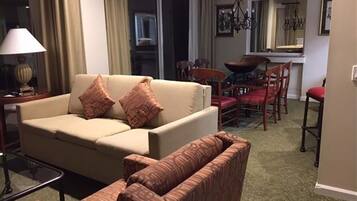 Un salon comprenant un canapé, deux fauteuils, une table à manger avec des chaises, un lustre et une vue sur la cuisine.