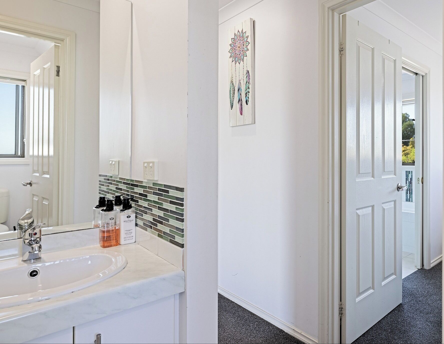 Een badkamer met een wit wastafelmeubel, een spiegel en een betegelde achterwand. Er is een deur naar een andere kamer.