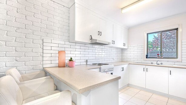 A modern kitchen with white cabinetry, a brick accent wall, a small island with seating, and a window with a view of greenery. - Regent Retreat - Sleeps 15, BBQ, pool (Moama)