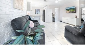 A modern living room with a grey sofa, a dining area, and a kitchen in the background. - Regent Retreat - Sleeps 15, BBQ, pool (Moama)