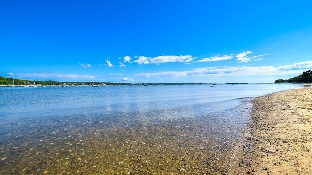 A clear view of a beach with calm waters, a sandy shore, and a distant shoreline with greenery and buildings. - Central AC Close to Beach Dog Friendly! (Chatham)