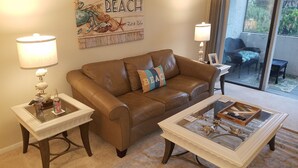 Una sala de estar con un sofá de cuero marrón, dos mesas auxiliares con lámparas, una mesa de centro con objetos decorativos y un cartel en la pared que dice 'Welcome to the Beach'.