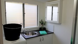 Et lille køkken med vask, et vindue med skodder, en sort spand, et grønt håndklæde og en potteplante.