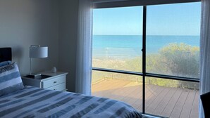 A bedroom with a bed, a nightstand, a lamp, a ceiling fan, and a large window with a view of the ocean.