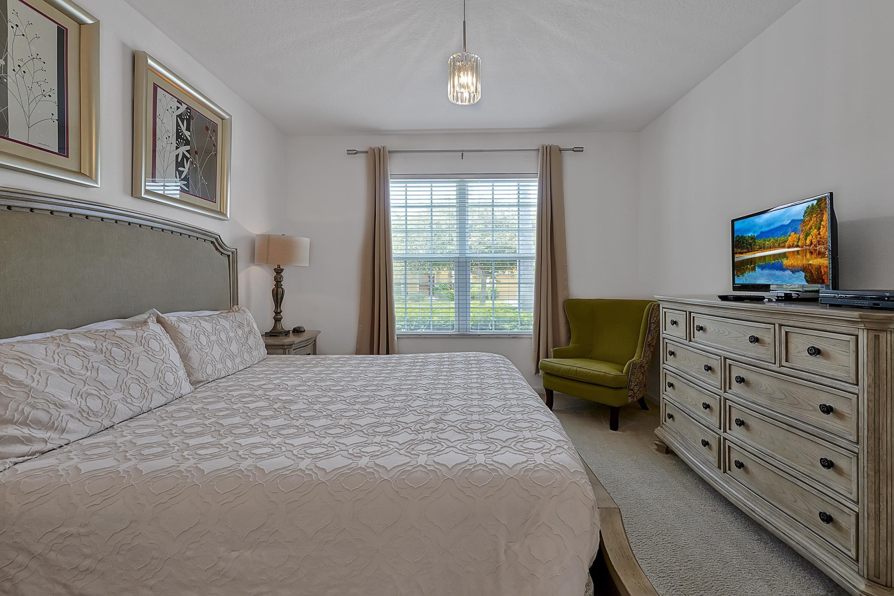 A neatly arranged bedroom with a large bed, a television, a green armchair, and a wooden dresser.