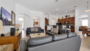 Amplia sala de estar con un sofá grande de color gris, una mesa de centro y una mesita de noche de madera. Se observa una zona de cocina con armarios de madera y un comedor con una silla de mimbre.