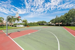 An outdoor basketball court with a red and green playing surface, surrounded by trees and a clear sky.