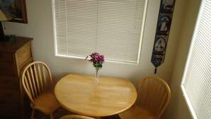 Un comedor con una mesa redonda de madera, dos sillas de madera, un jarrón con flores moradas, una lámpara, un aparador de madera y una ventana con persianas.
