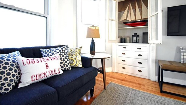 A cozy living room with a dark blue sofa, a small round table with a lamp, and a white cabinet with drawers. - Convenient, Modern Thompson s Point 3-Bed Flat (Portland)