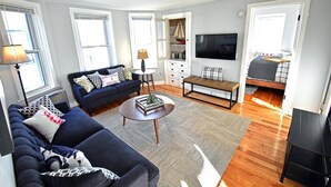 A modern living room with a sofa, coffee table, and a TV. There are windows, a bookshelf, and a bench. - Convenient, Modern Thompson s Point 3-Bed Flat (Portland)