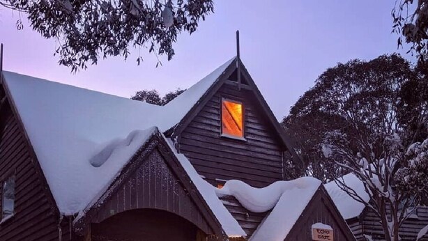 A wooden cabin with a snow-covered roof and a lit interior, surrounded by snow. - Echo Base – Alpine Family Retreat, Dinner Plain (Dinner Plain)