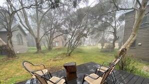 A wooden deck with two wicker chairs and a table, surrounded by trees and houses in a foggy setting. - Echo Base – Alpine Family Retreat, Dinner Plain (Dinner Plain)