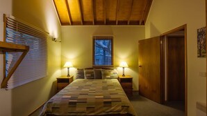 A bedroom with a wooden ceiling, a bed with a quilt, two bedside lamps, a window with blinds, and a door. - Echo Base – Alpine Family Retreat, Dinner Plain (Dinner Plain)