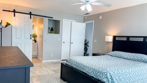 Un dormitorio con cama, escritorio, ventilador de techo y puerta que da a otra habitación.