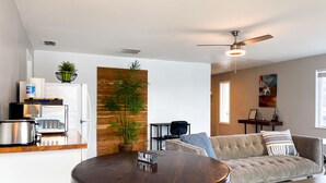 Una sala de estar moderna con una mesa de centro redonda de madera, un sofá beige, un ventilador de techo y una zona de cocina con microondas y tostadora.