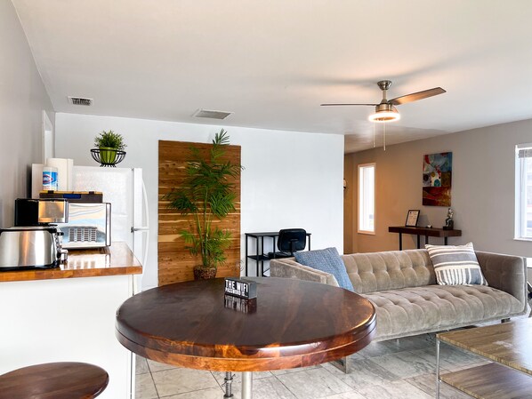 A modern living room with a round wooden coffee table, a beige sofa, a ceiling fan, and a kitchen area with a microwave and toaster.