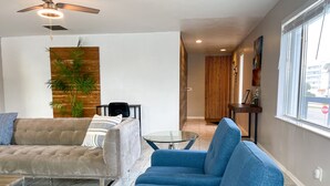 Una sala de estar moderna con un sofá, un sillón azul, una mesita de centro de vidrio y un ventilador de techo.