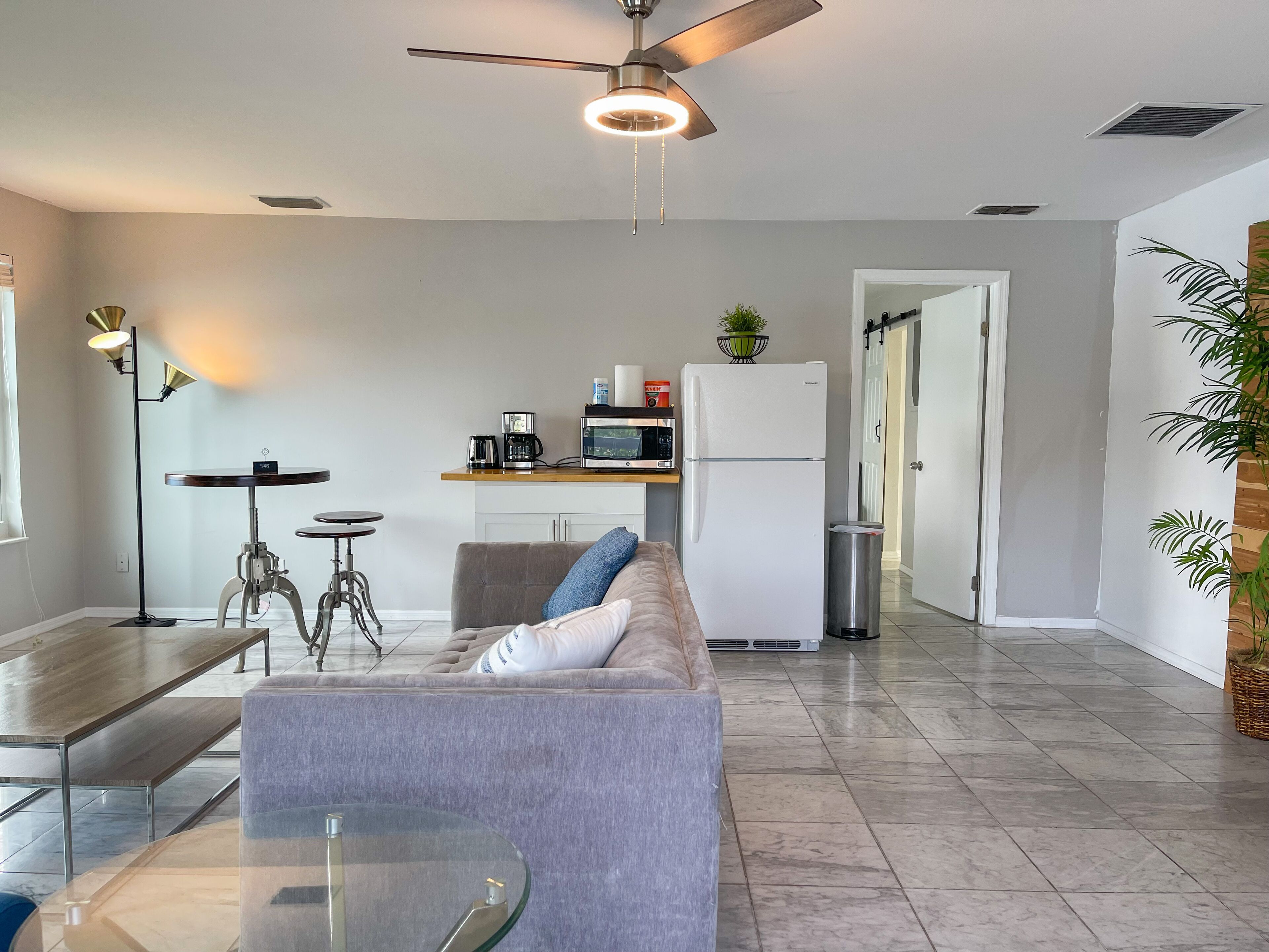 A modern living room with a sofa, coffee table, and a dining area with a table and chairs. A kitchen area with a refrigerator, microwave, and coffee maker is visible in the background.
