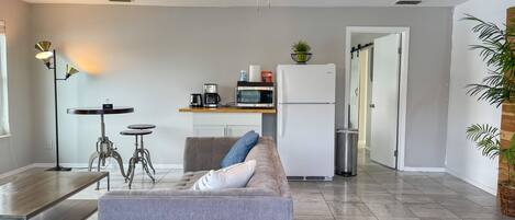 A modern living room with a sofa, coffee table, and a dining area with a table and chairs. A kitchen area with a refrigerator, microwave, and coffee maker is visible in the background.