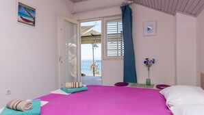 Ein Schlafzimmer mit Bett, Fenster mit Jalousien, einer Tür zu einem Außenbereich, einem kleinen Tisch mit Blumenvase und einem Bild an der Wand.