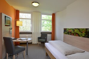 A hotel room with a bed, a desk with a chair, a television, and a window with curtains. - Tagungszentrum Schmerlenbach (Hoesbach)