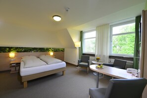 A hotel room with a bed, a desk, a chair, a table with a fruit bowl, and a window with curtains. - Tagungszentrum Schmerlenbach (Hoesbach)