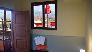 Una habitación con cama, una silla, un ventanal con vistas a casas y montañas, y una puerta que da a un espacio al aire libre.