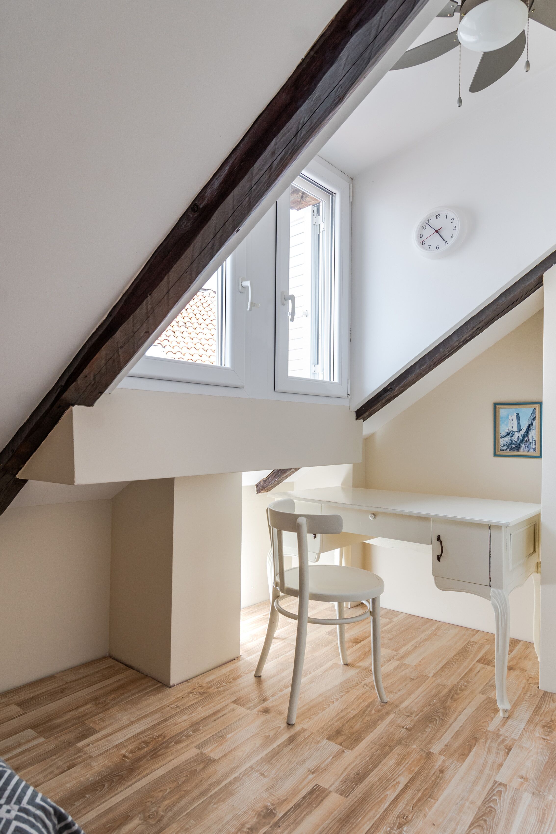 A room with a sloped ceiling, a desk, a chair, a clock, and a window.