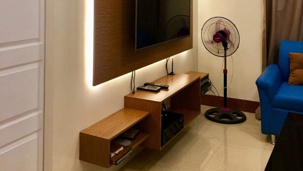 Un salon moderne avec un téléviseur à écran plat fixé sur un panneau en bois, un ventilateur sur pied, un canapé bleu et une petite table.