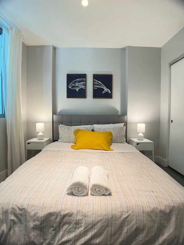 A neatly made bed with a striped bedspread, two pillows, and two rolled towels. There are two bedside tables with lamps, a headboard, and two framed wall art pieces. - Luxurious 2BR, Pool, Hot tub, Sauna, Gym, Parking, Min to Beach (Miami Beach)
