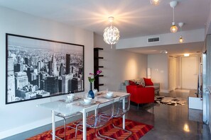 A modern living room with a dining area, featuring a cityscape artwork, a glass dining table, clear chairs, a red sofa, and a chandelier. - Luxurious 2BR, Pool, Hot tub, Sauna, Gym, Parking, Min to Beach (Miami Beach)
