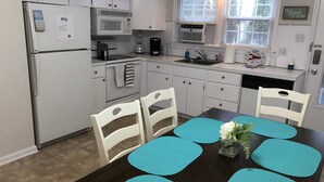 Een keuken met witte kasten, een koelkast, een magnetron, een fornuis en een spoelbak. Er staat een eettafel met stoelen en blauwe placemats.