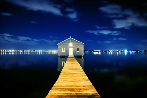 A wooden pier leading to a small, light-colored house with a gabled roof, situated on calm water with a dark night sky. - Balcony, bath, kitchen, laundry, pool, tennis court, gym, designated car bay (Perth)