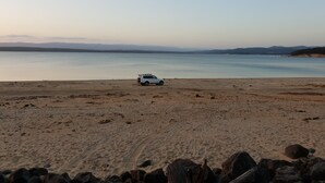 Um SUV branco estacionado numa praia arenosa, com águas calmas e montanhas ao longe.