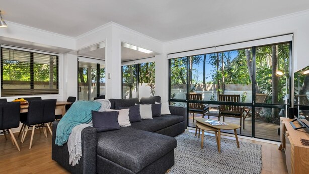 A modern living room with a grey sofa, black chairs, and a wooden coffee table. Large windows offer a view of the outdoors. - Mahogany Lodge - townhouse near Tallow Beach (Byron Bay)