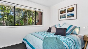A bedroom with a large window, a bed with patterned bedding, a nightstand, and a framed picture on the wall. - Mahogany Lodge - townhouse near Tallow Beach (Byron Bay)