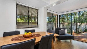 A modern dining area with a wooden table, black chairs, and a bowl of fruit. Large windows with blinds provide views of the outdoors. - Mahogany Lodge - townhouse near Tallow Beach (Byron Bay)