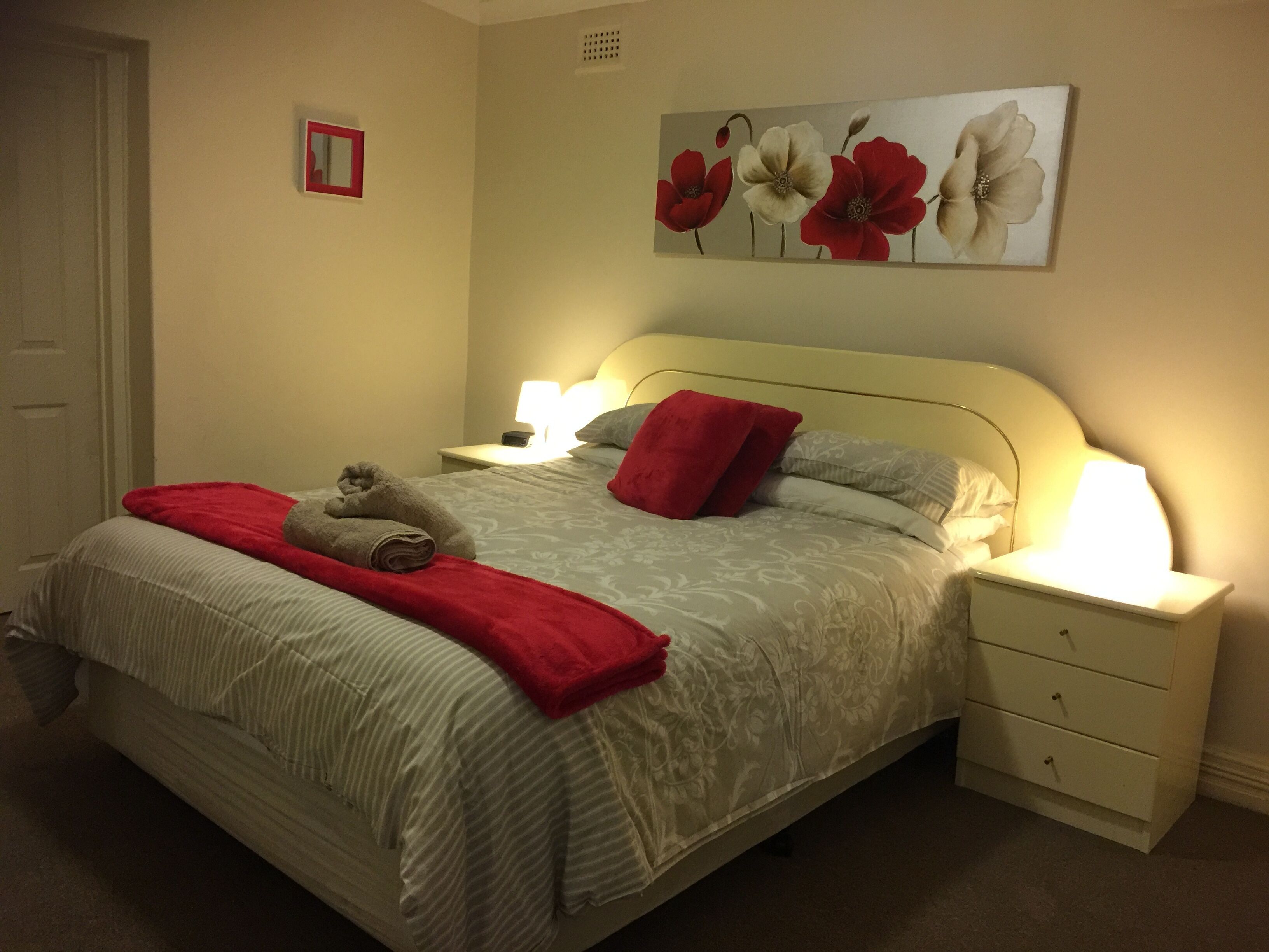Un lit bien fait, avec une couverture rouge et des oreillers, une table de chevet avec une lampe, et un tableau de fleurs accroché au mur.