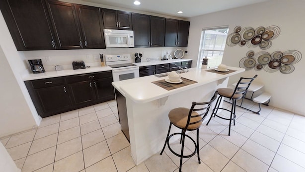 A modern kitchen with dark cabinets, a white countertop, a central island with stools, and a decorative wall piece. - Affordable 4 bedrooms at Crystal Cove (1527) (Kissimmee)