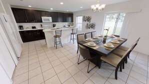 A modern kitchen with dark cabinets, a white island, and a dining area with a large table set for six. - Affordable 4 bedrooms at Crystal Cove (1527) (Kissimmee)