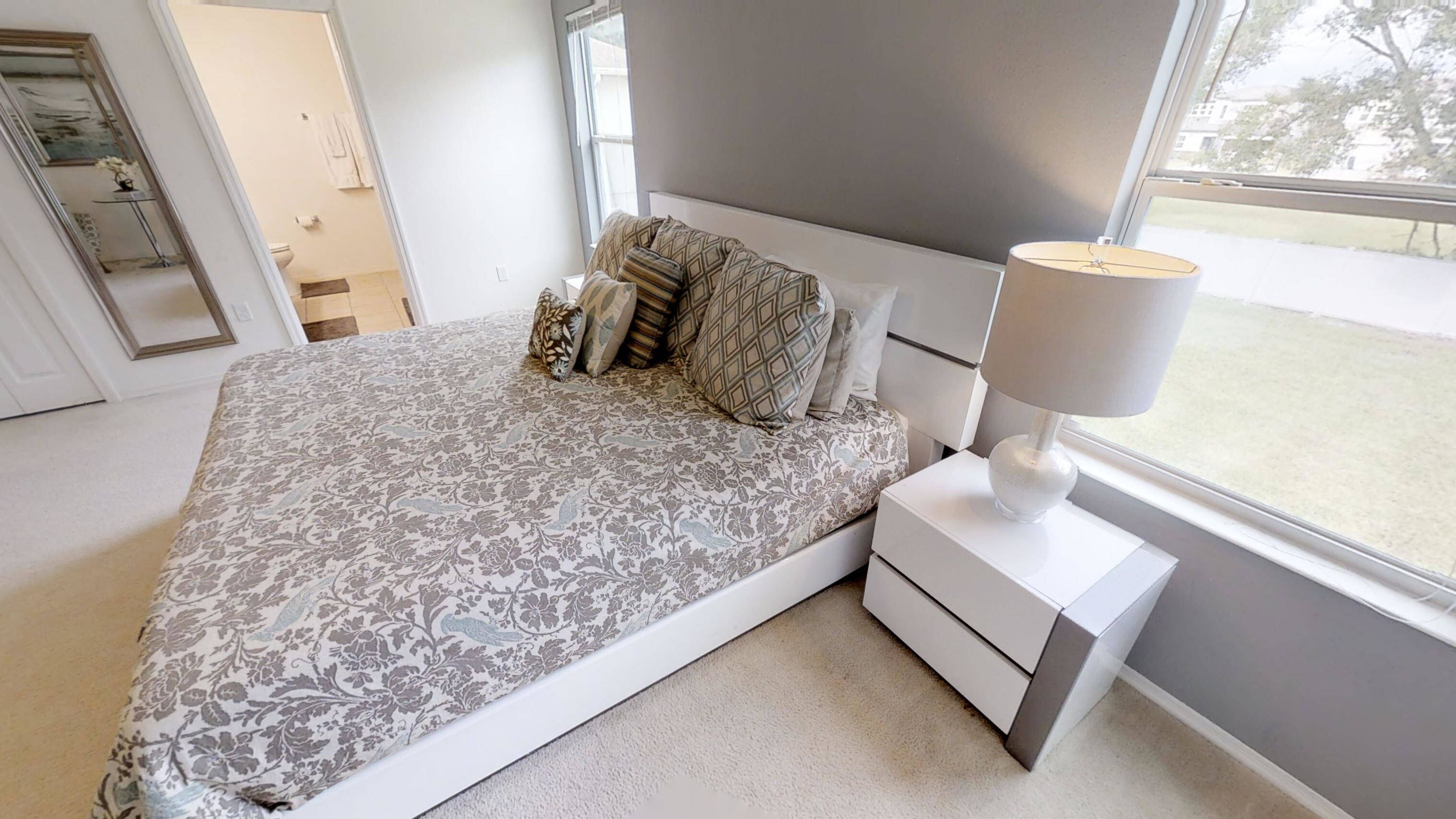 A modern bedroom with a large bed, a nightstand, a lamp, and a view of the outside.
