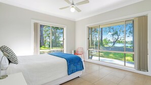Une chambre à coucher avec un lit, un ventilateur de plafond et de grandes fenêtres donnant sur des arbres et une route.