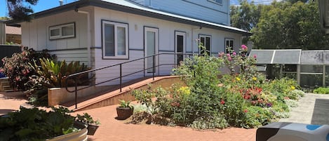A two-story building with a white exterior and black trim, a covered outdoor area with a metal railing, and a variety of potted plants and flowers.