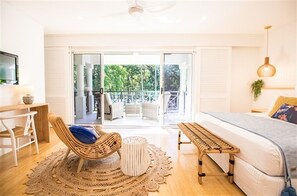 Relax in this serene bedroom with a large bed, wooden bench, and wicker chair. Enjoy the view from the sliding glass door leading to a balcony with outdoor seating.