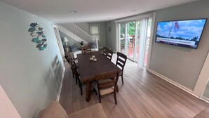 A dining area with a wooden table and chairs, a staircase leading to another level, and a television mounted on the wall displaying a news broadcast.
